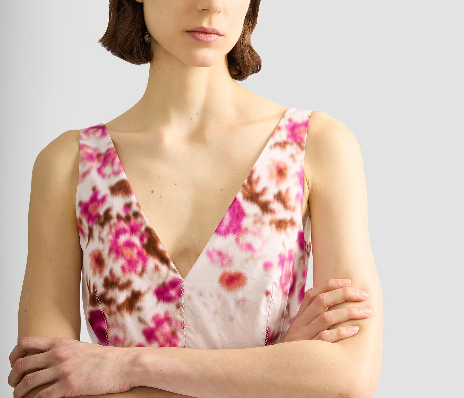 A woman with short brown hair stands with arms crossed, wearing the Erdem Rose Bouquet Fit and Flared Midi Dress—a sleeveless cotton poplin dress featuring a pink and brown floral pattern—against a plain background.