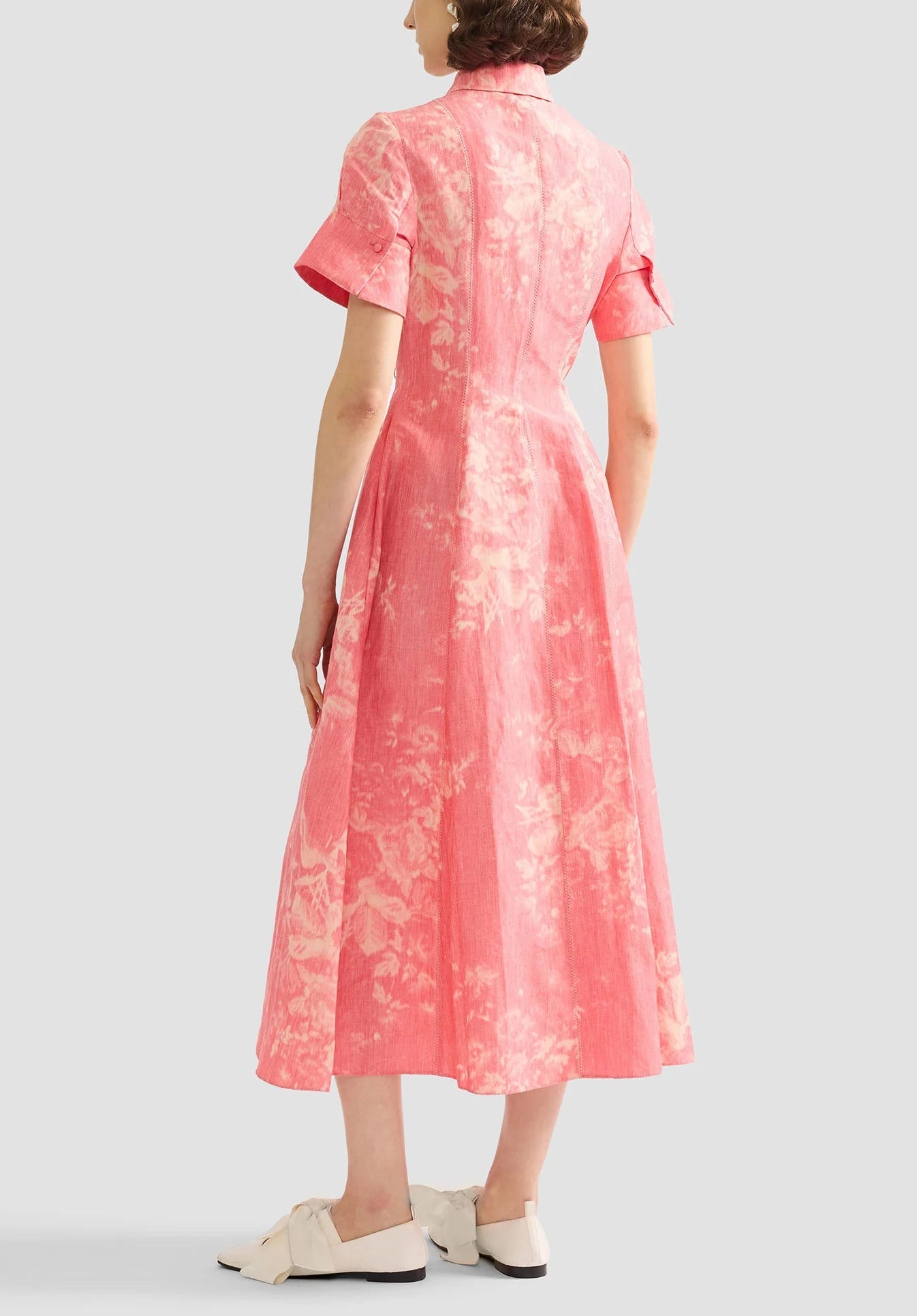 A woman faces away, wearing the Erdem Rose Bouquet Linen Chine Midi Shirt Dress with short sleeves and white bow slip-ons, set against a plain light background.