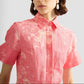 A woman wears the Erdem Rose Bouquet Linen Chine Midi Shirt Dress, featuring a rose-pink hue and short sleeves, paired with pearl drop earrings. The image is cropped from her shoulders to mid-chest.