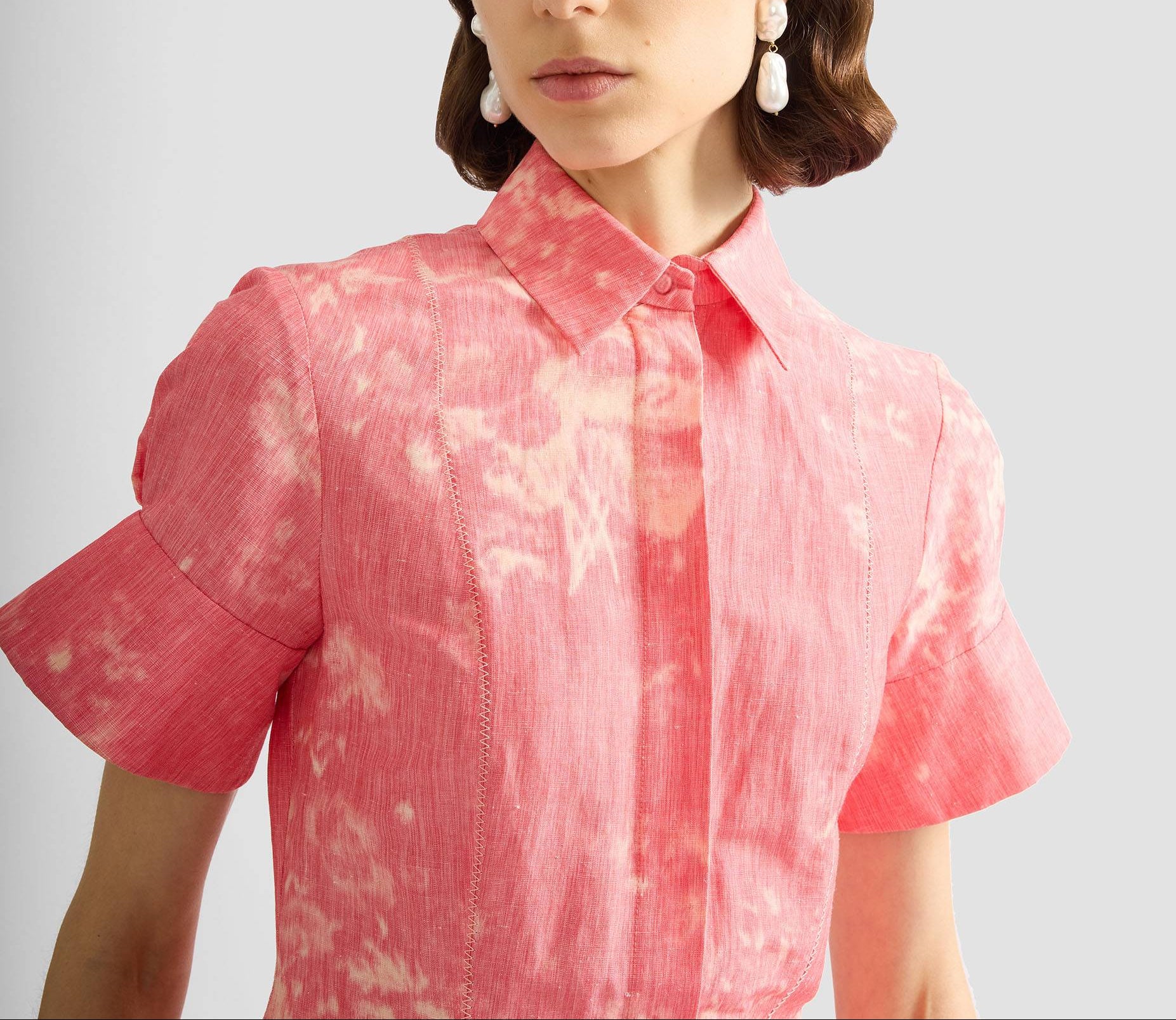 A woman wears the Erdem Rose Bouquet Linen Chine Midi Shirt Dress, featuring a rose-pink hue and short sleeves, paired with pearl drop earrings. The image is cropped from her shoulders to mid-chest.