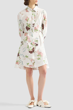 A person wears the Erdem French Chintz Mini Shirt Dress, a white floral cotton poplin button-down with long sleeves, paired with cream bow-accented shoes, standing against a light gray background.