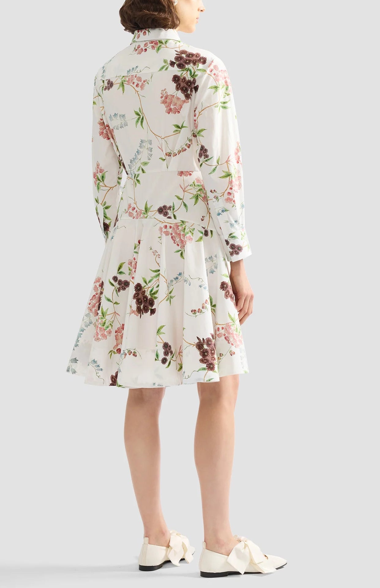 A person stands facing away, wearing the Erdem French Chintz Mini Shirt Dress with a floral pattern and white shoes featuring large bows.