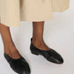 A person in a beige skirt wears Mansur Gavriel's Ruched Ballerina flats in black Italian leather, standing on a light surface.