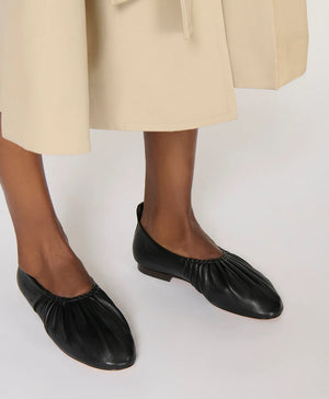 A person in a beige skirt wears Mansur Gavriel's Ruched Ballerina flats in black Italian leather, standing on a light surface.