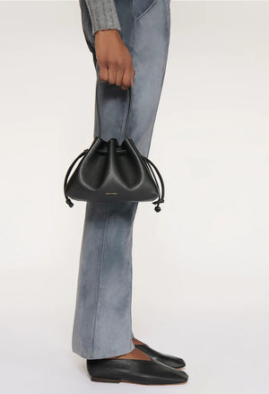 A person in gray pants and black shoes holds the Mansur Gavriel Drawstring Pouchette, a small black Italian leather handbag, by its handles against a plain background.