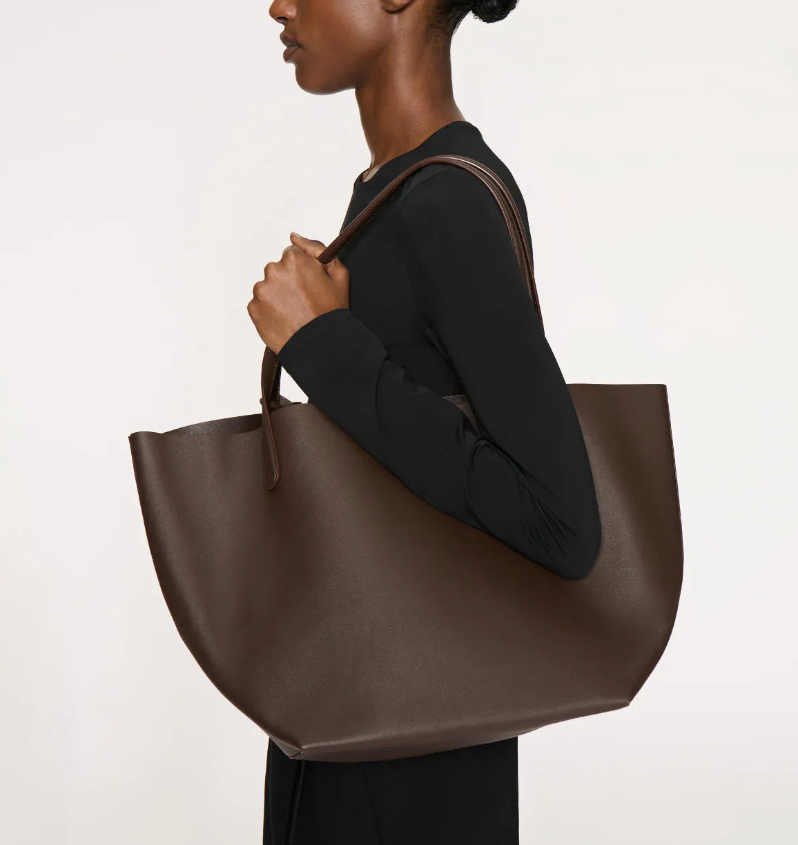 Dressed in black, a person carries the spacious Everyday Soft Tote by Mansur Gavriel over their shoulder against a plain background.
