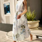 A woman in a Monique Lhuillier Wisteria Floral Poplin Day Dress with an A-line skirt and clear heels walks outside, passing a large planter and a building with tall vertical panels.