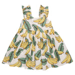 The Girls' Via Dress by Pink Chicken is a white sleeveless cotton cambric dress with ruffled, adjustable straps and a playful tropical banana and green leaf print.