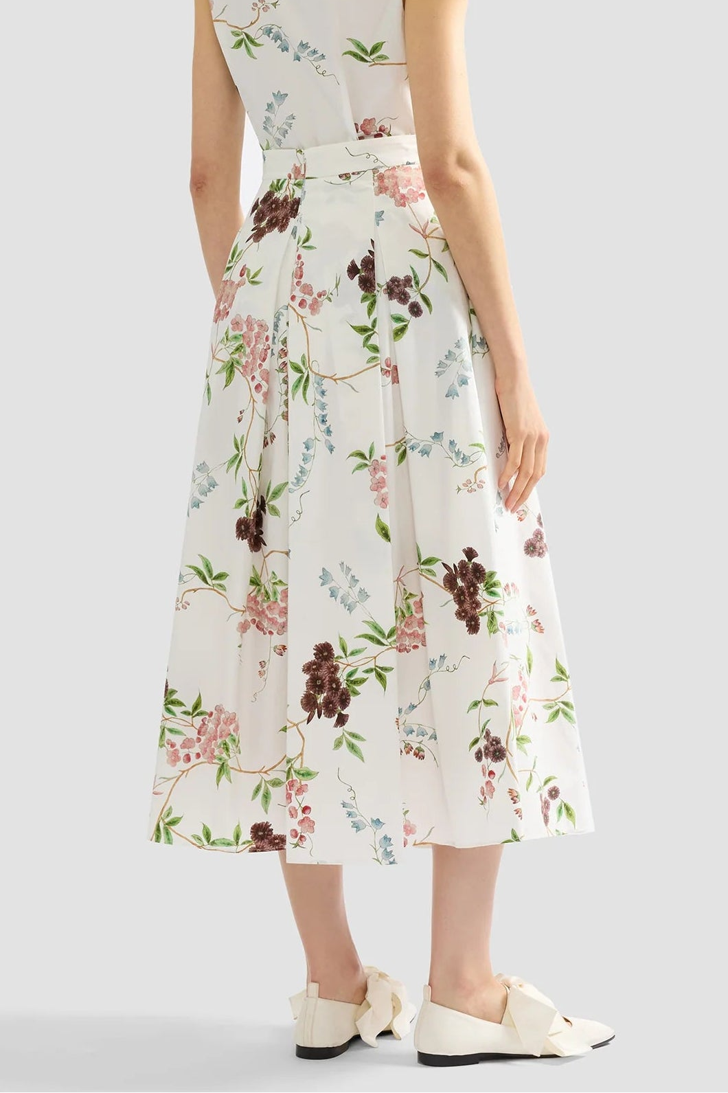 A person stands against a plain background wearing Erdem’s French Chintz Cotton Midi Skirt, styled with white flat shoes featuring bow details.