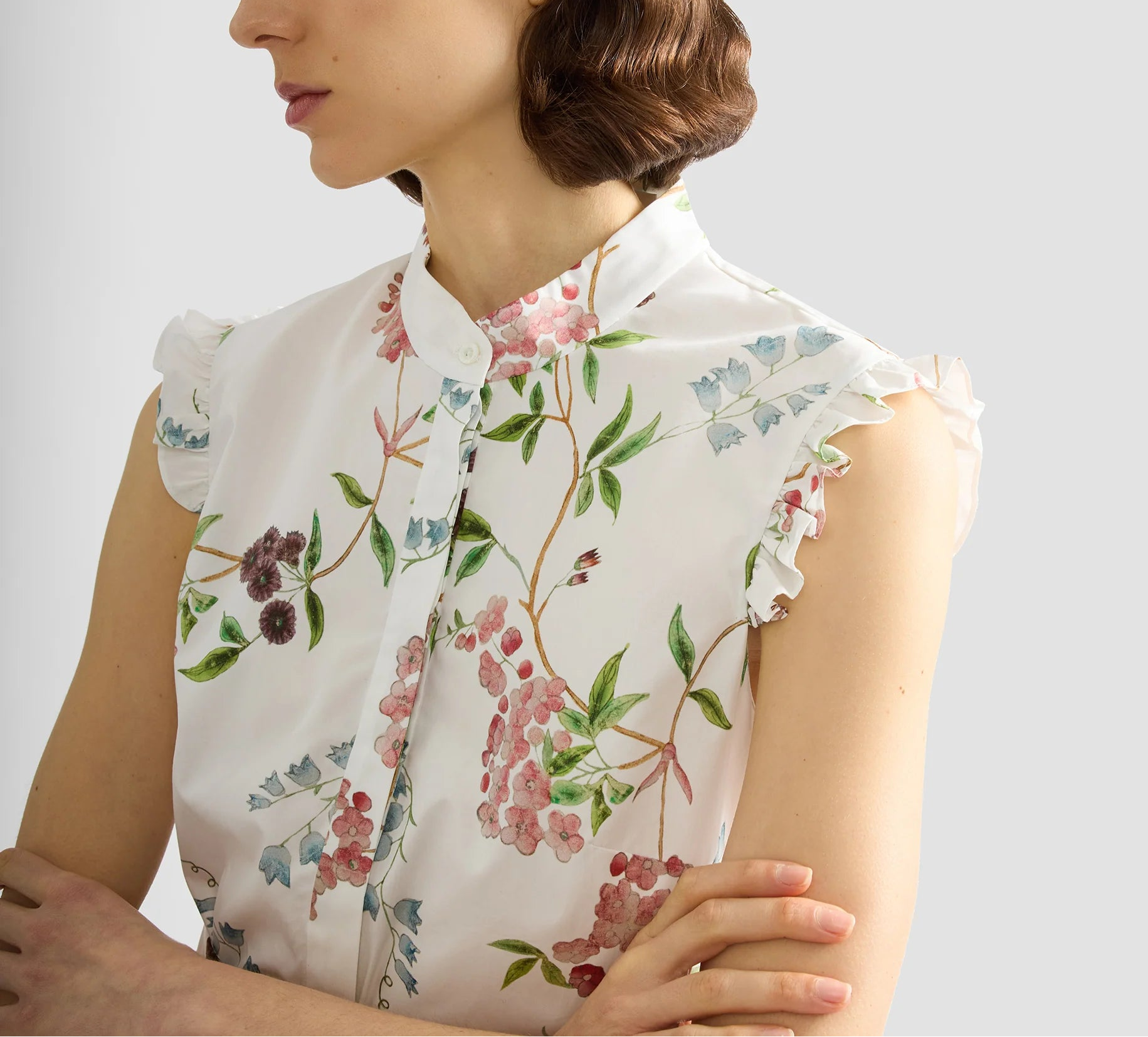 A woman in the Erdem French Chintz Cotton Sleeveless Shirt, featuring a white floral print and ruffle trim, stands with her arms crossed.