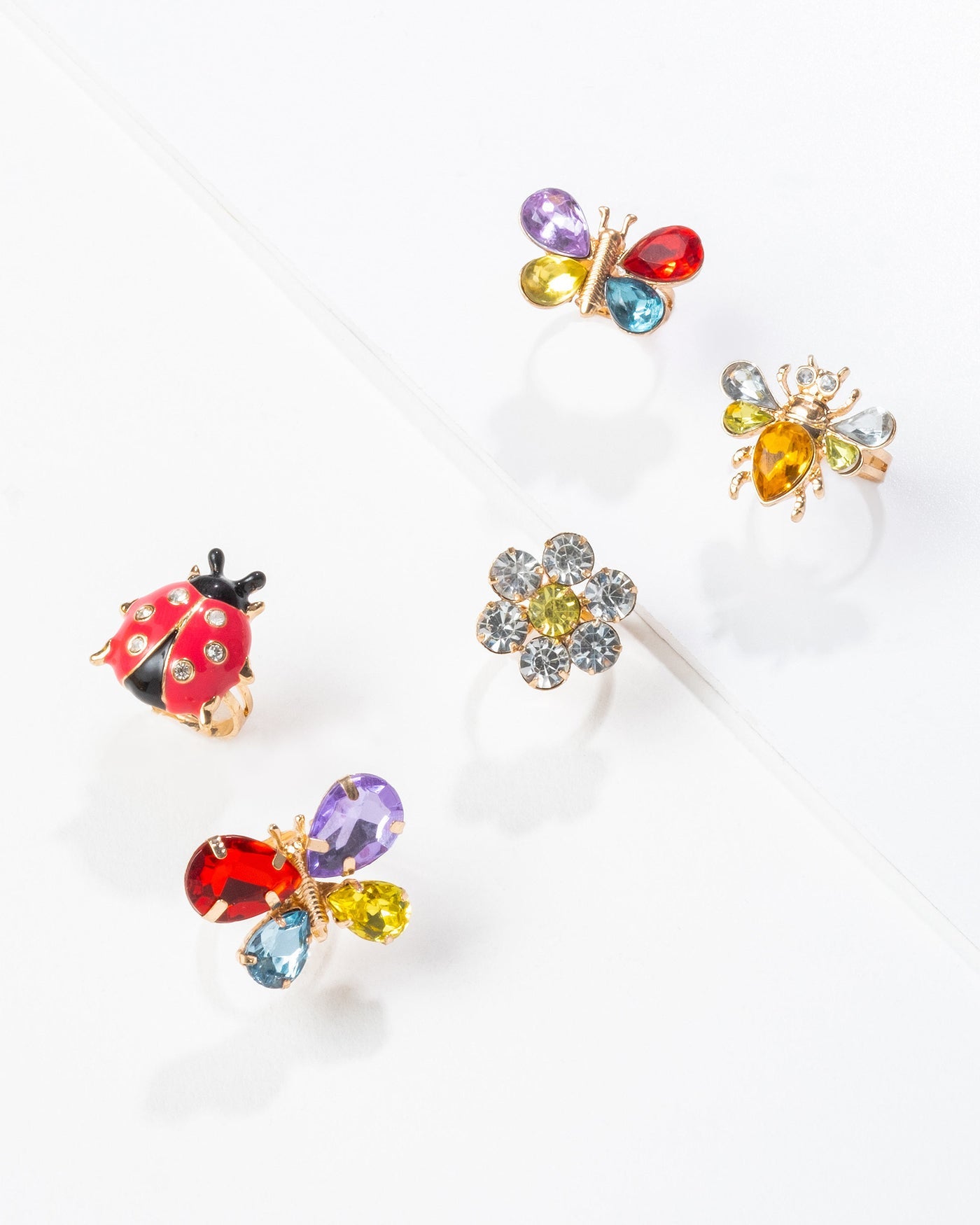 The Super Smalls Garden Get-Together Ring Set features five playful kids rings—a ladybug, a bee, a flower, and two colorful butterfly designs—arranged on a white surface and adorned with vibrant gemstones.