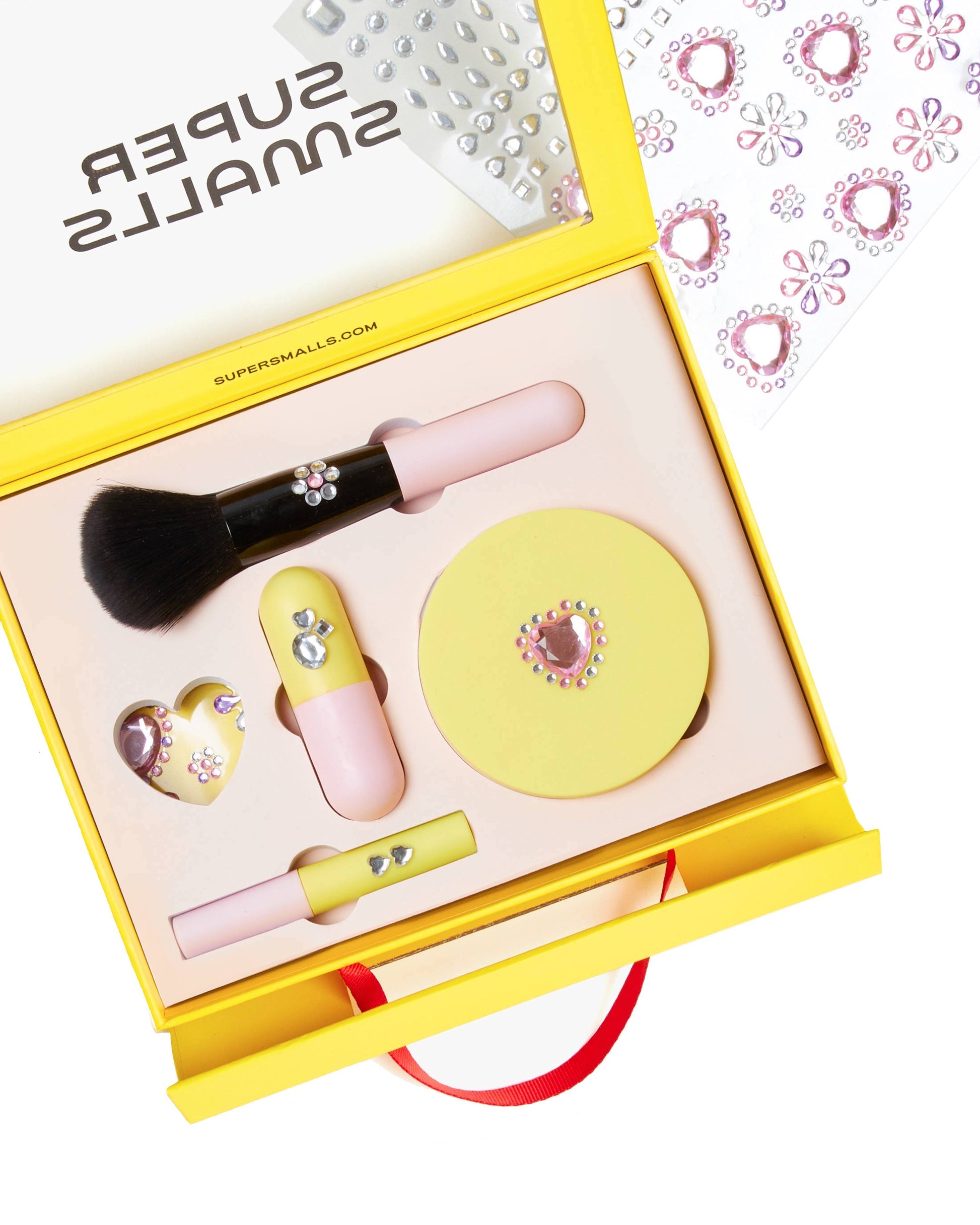 The Super Smalls Mom's Makeup Play Kit is a yellow kids set with brushes, lip products, a compact, nail accessories, and rhinestones. A sheet of rhinestone stickers appears in the background. Safe makeup designed for children to enjoy.