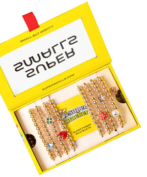 A yellow box labeled "Super Stack Bracelet Set" by Super Smalls holds gold beaded bracelets with colorful charms, perfect for mixing, matching, or everyday accessorizing.