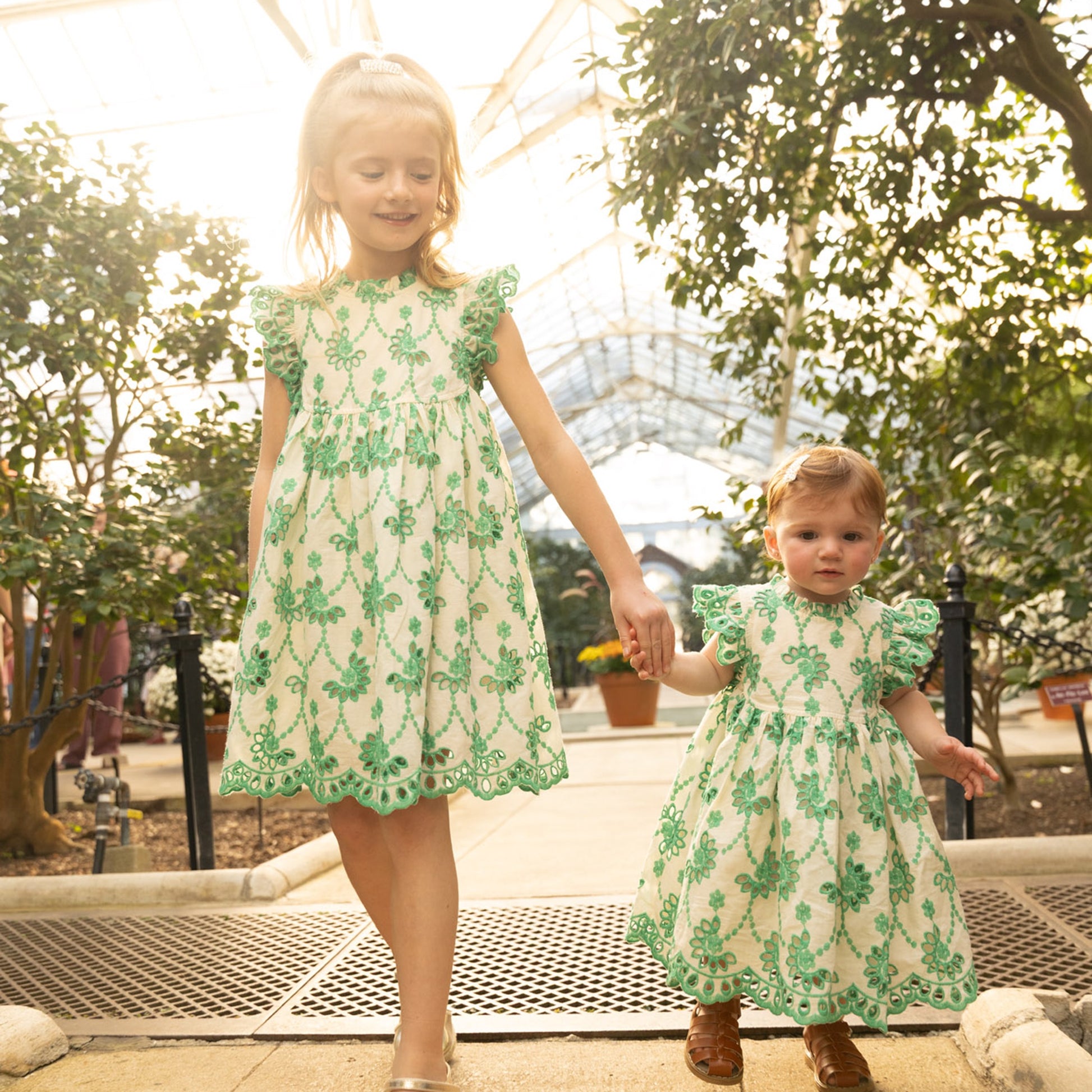 Two young girls hold hands in a greenhouse, both dressed in matching Pink Chicken Baby Girls' Cynthia Dresses—white with green floral patterns.
