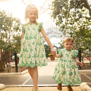 Two young girls hold hands in a greenhouse, both dressed in matching Pink Chicken Baby Girls' Cynthia Dresses—white with green floral patterns.