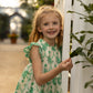 A young girl smiles outdoors next to a white structure and greenery, wearing the Pink Chicken Girls' Cynthia Dress, featuring blonde hair and a green eyelet design.