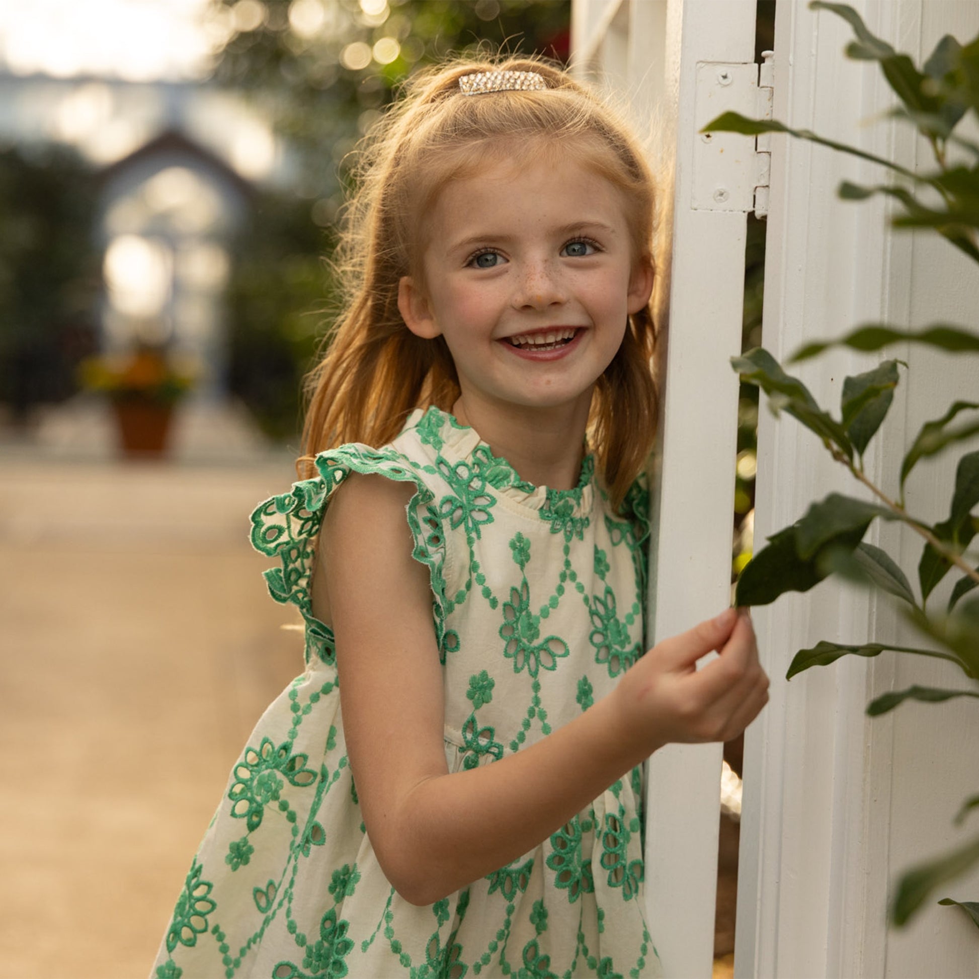 A young girl smiles outdoors next to a white structure and greenery, wearing the Pink Chicken Girls' Cynthia Dress, featuring blonde hair and a green eyelet design.