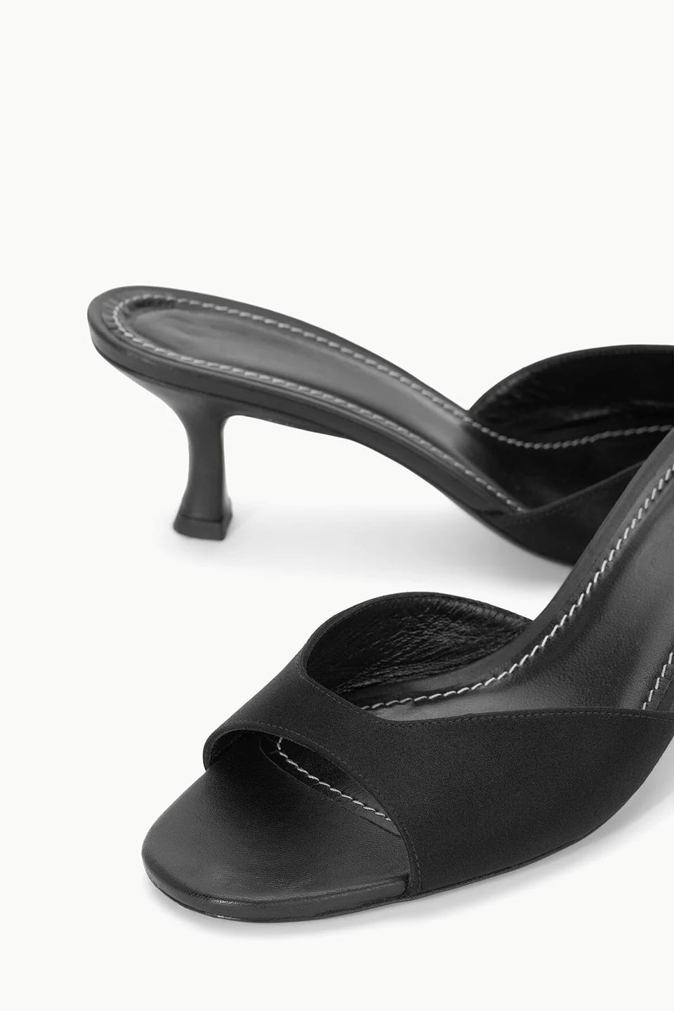 Close-up of the Brigitte Satin Mule by Staud, a black open-toe mule with a satin kitten heel and white stitching along the insole—ideal for versatile styling.