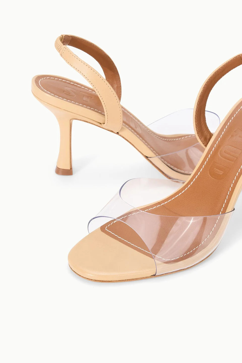 The Staud Brigitte Heel is a beige sandal featuring clear PVC straps, a slim high heel, and a chic slingback design, shown against a white background.