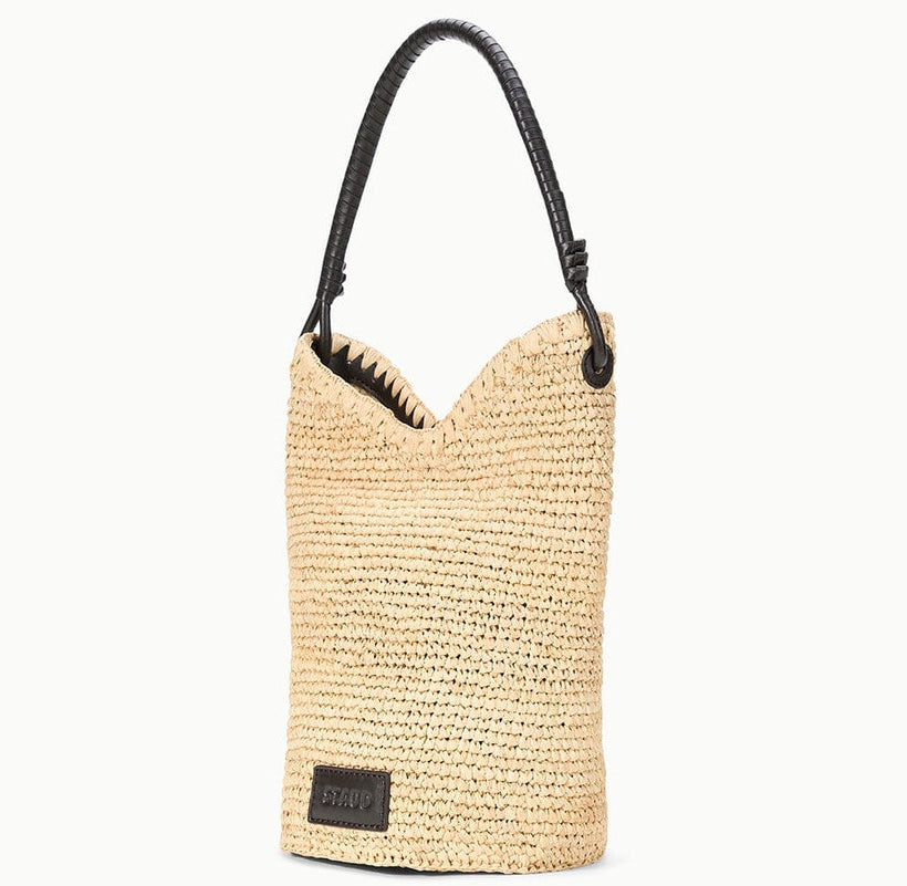 The Lani Raffia Bucket Bag by Staud features beige raffia, a black leather handle, and a black rectangular brand label—an everyday essential for effortless style.