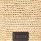 Close-up of the Staud Lani Raffia Bucket Bag, showcasing its woven straw design, rectangular black leather patch with embossed branding, and sturdy leather handle—a stylish yet practical everyday accessory.