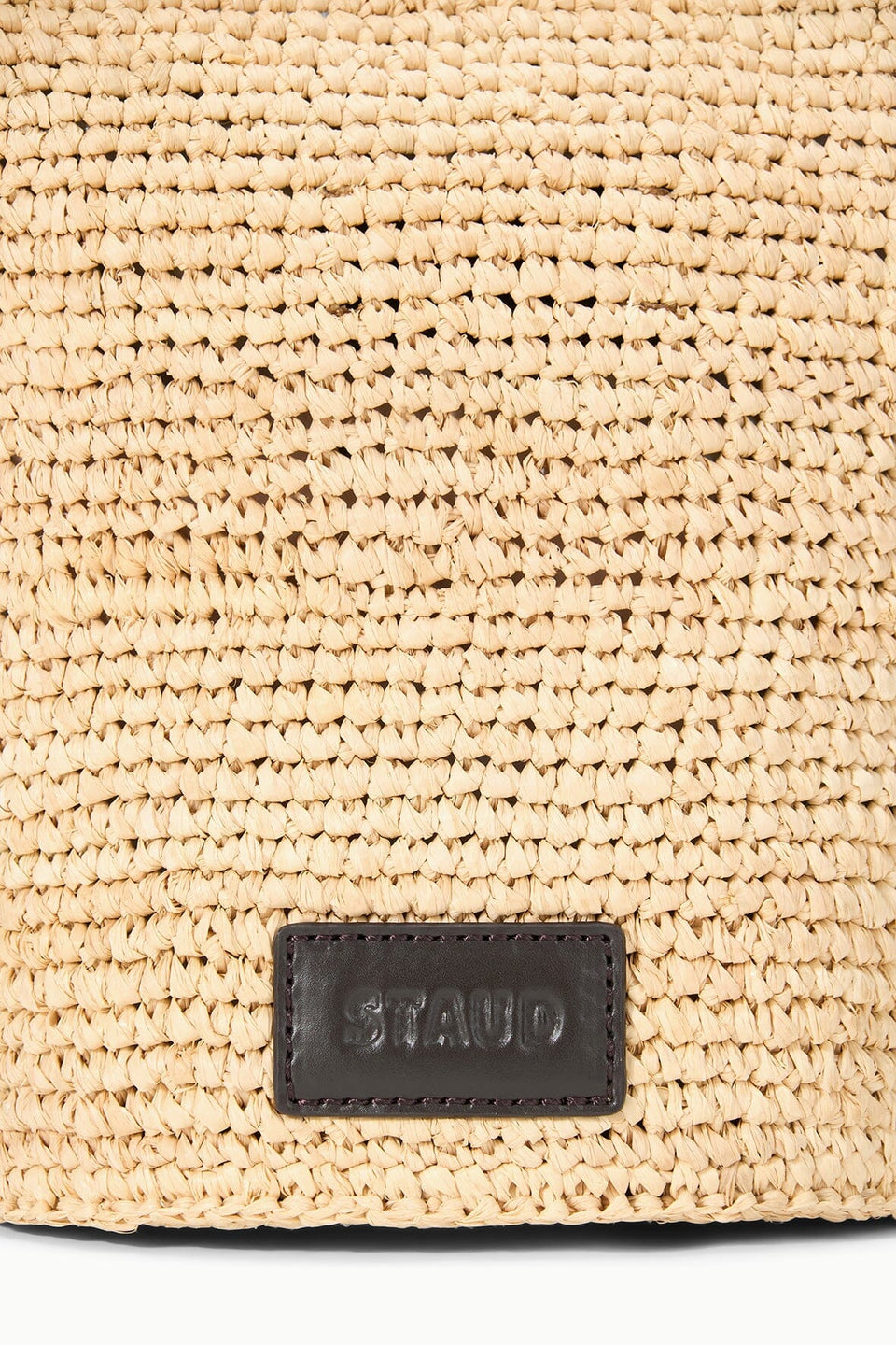 Close-up of the Staud Lani Raffia Bucket Bag, showcasing its woven straw design, rectangular black leather patch with embossed branding, and sturdy leather handle—a stylish yet practical everyday accessory.