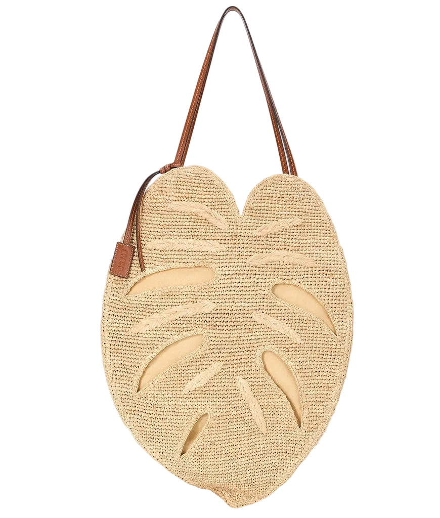 The Staud Large Palm Raffia Bag is a unique woven carryall, shaped like a monstera leaf with brown shoulder straps and tan accents, shown against a white background.