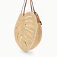 The Staud Large Palm Raffia Bag features a round, monstera leaf-inspired silhouette with intricate cut-out details and two brown leather shoulder straps. Photographed on a white background, this spacious carryall tote combines natural woven raffia with sophisticated accents.