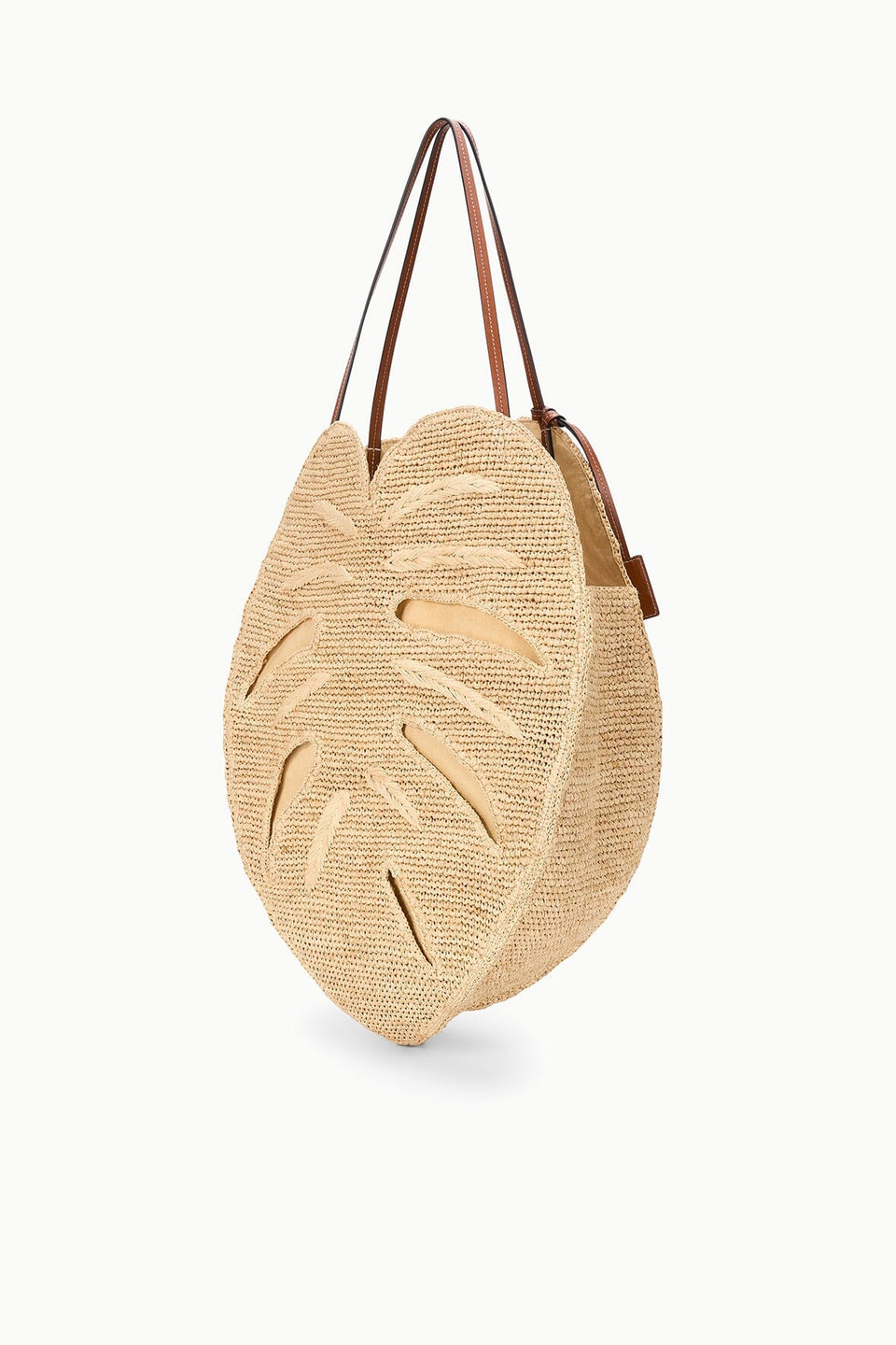 The Staud Large Palm Raffia Bag features a round, monstera leaf-inspired silhouette with intricate cut-out details and two brown leather shoulder straps. Photographed on a white background, this spacious carryall tote combines natural woven raffia with sophisticated accents.