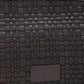 A close-up of the Staud Maude Woven Soft Clutch in dark brown leather, showcasing a rectangular patch with embossed text.