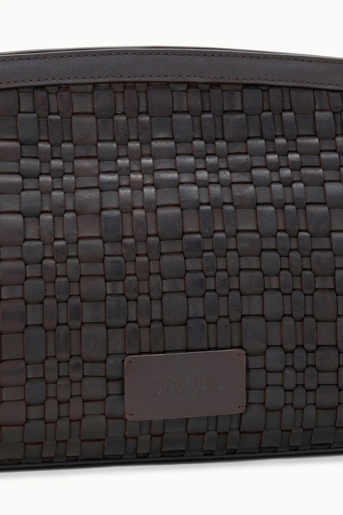 A close-up of the Staud Maude Woven Soft Clutch in dark brown leather, showcasing a rectangular patch with embossed text.