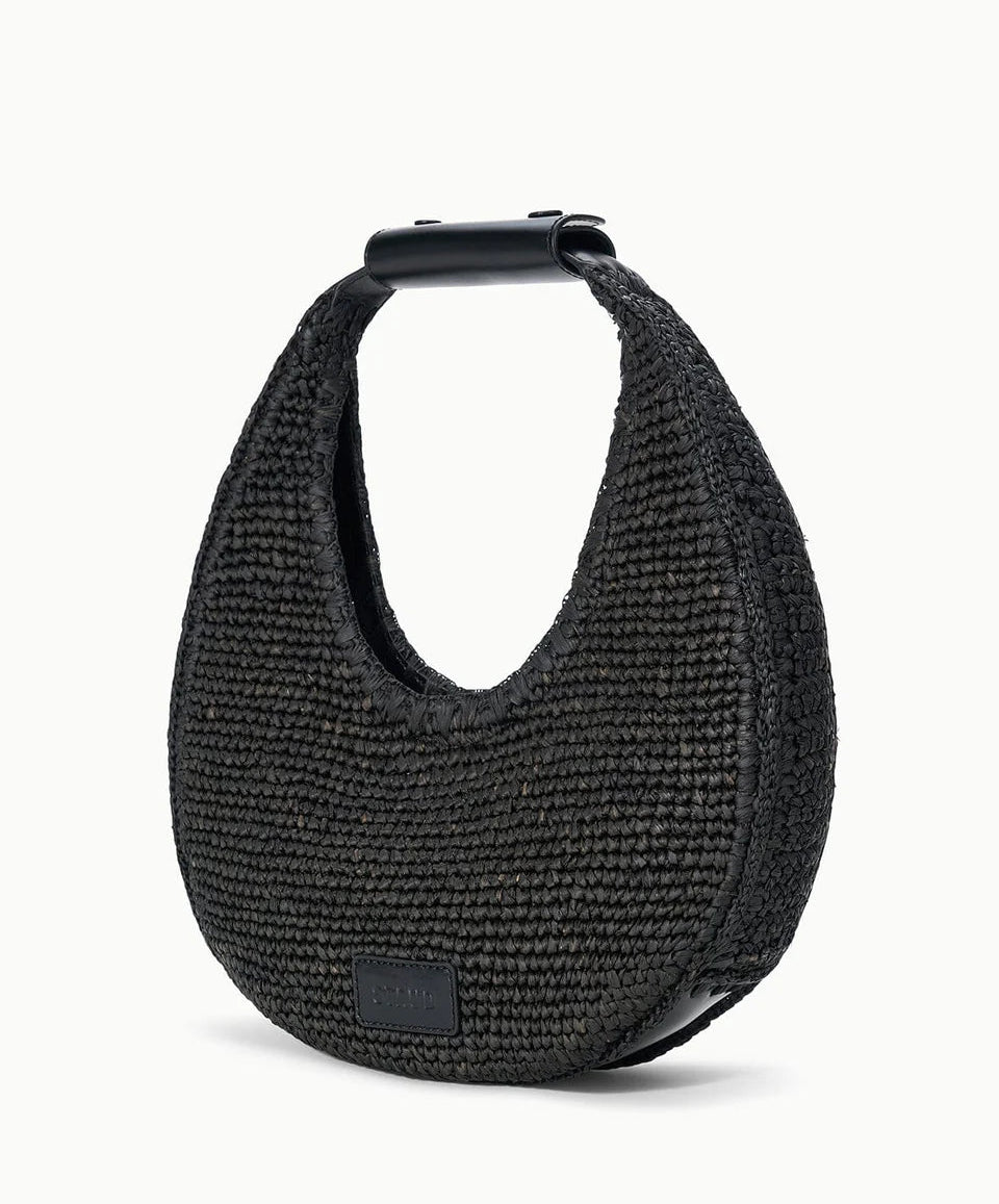 The Staud Moon Raffia Tote Bag is a black, crescent-shaped woven handbag with a short structured handle, magnetic closure, and a small rectangular logo patch on the lower front.