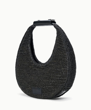 The Staud Moon Raffia Tote Bag is a black, crescent-shaped woven handbag with a short structured handle, magnetic closure, and a small rectangular logo patch on the lower front.