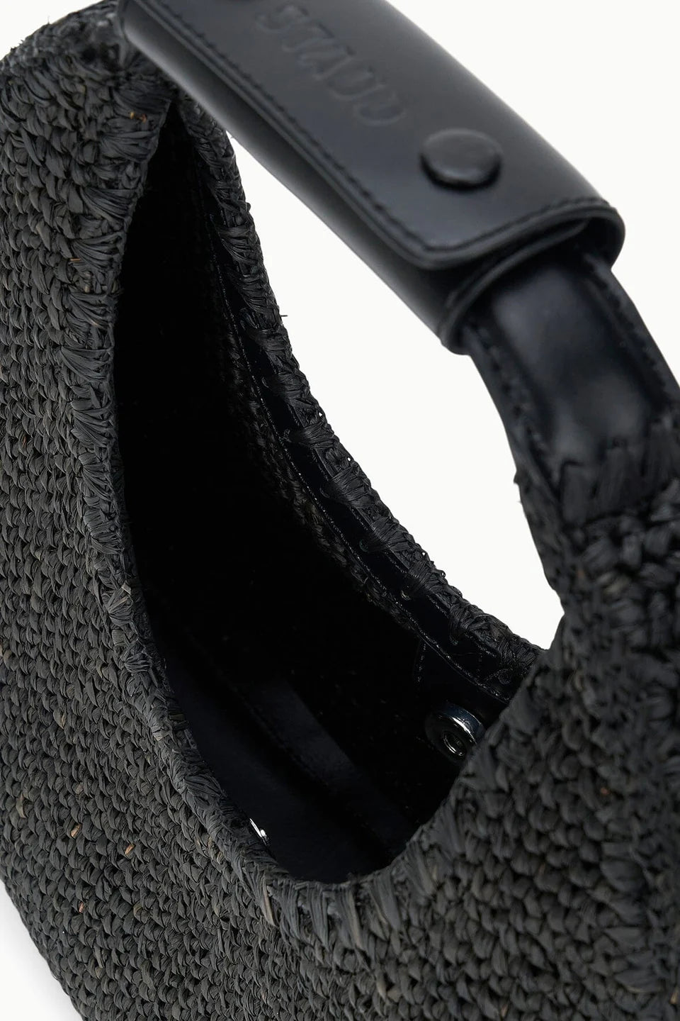 Close-up of the Staud Moon Raffia Tote Bag, featuring a black woven raffia body, textured surface, smooth black leather handle with visible stitching, snap detail, and a secure magnetic closure.
