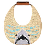 The Staud Moon Raffia Tote Bag features a brown handle and an embroidered shark head with blue waves, making it a playful and stylish choice for the beach.