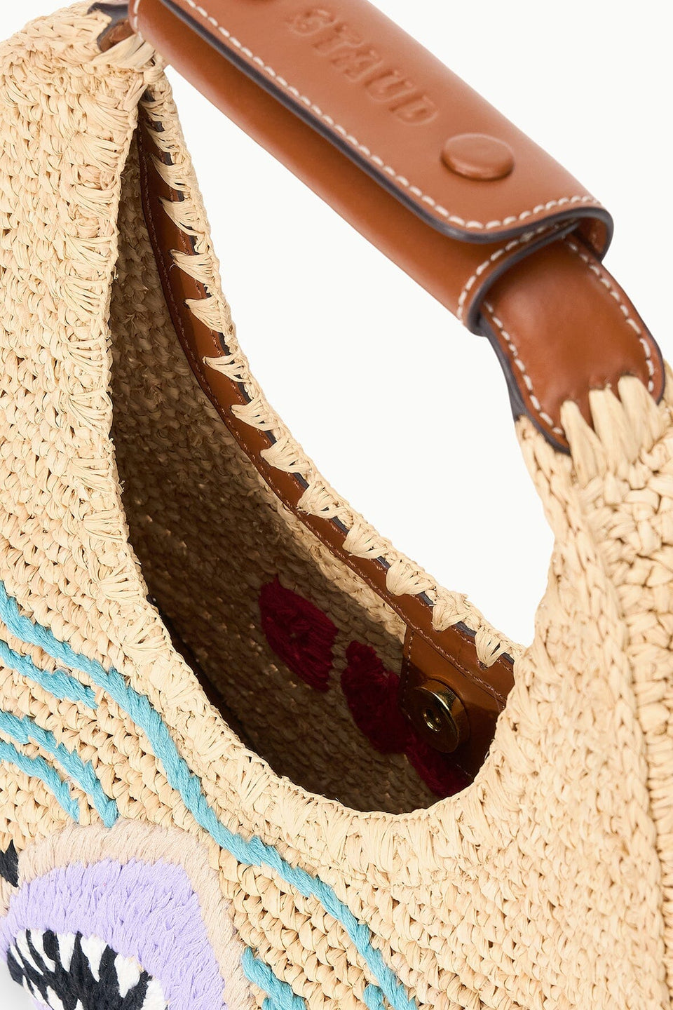 Close-up of the Staud Moon Raffia Tote Bag, featuring a brown leather handle, visible stitching, and colorful embroidered patterns on the exterior.