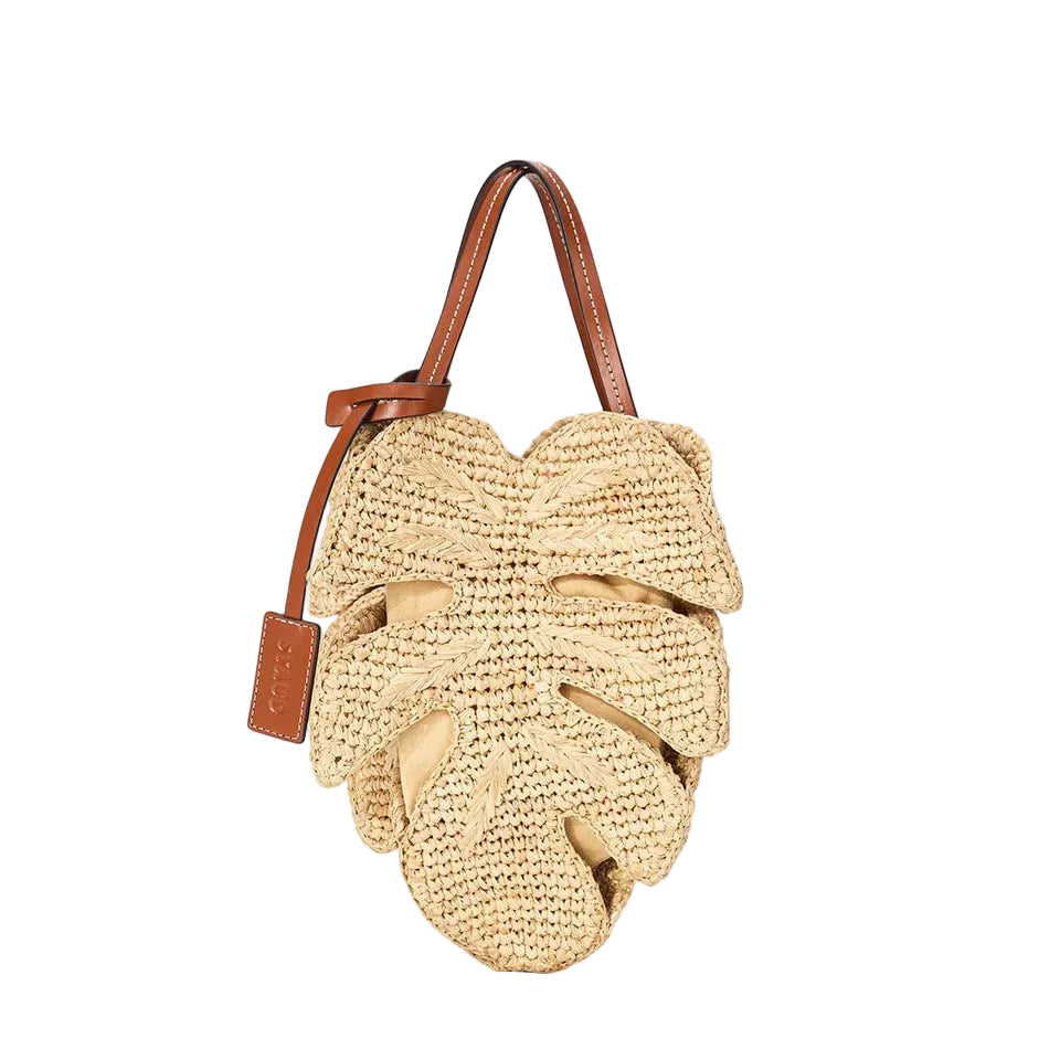 The Palm Raffia Bag by Staud is a woven raffia handbag shaped like a monstera leaf, featuring a tan leather strap and matching tag.