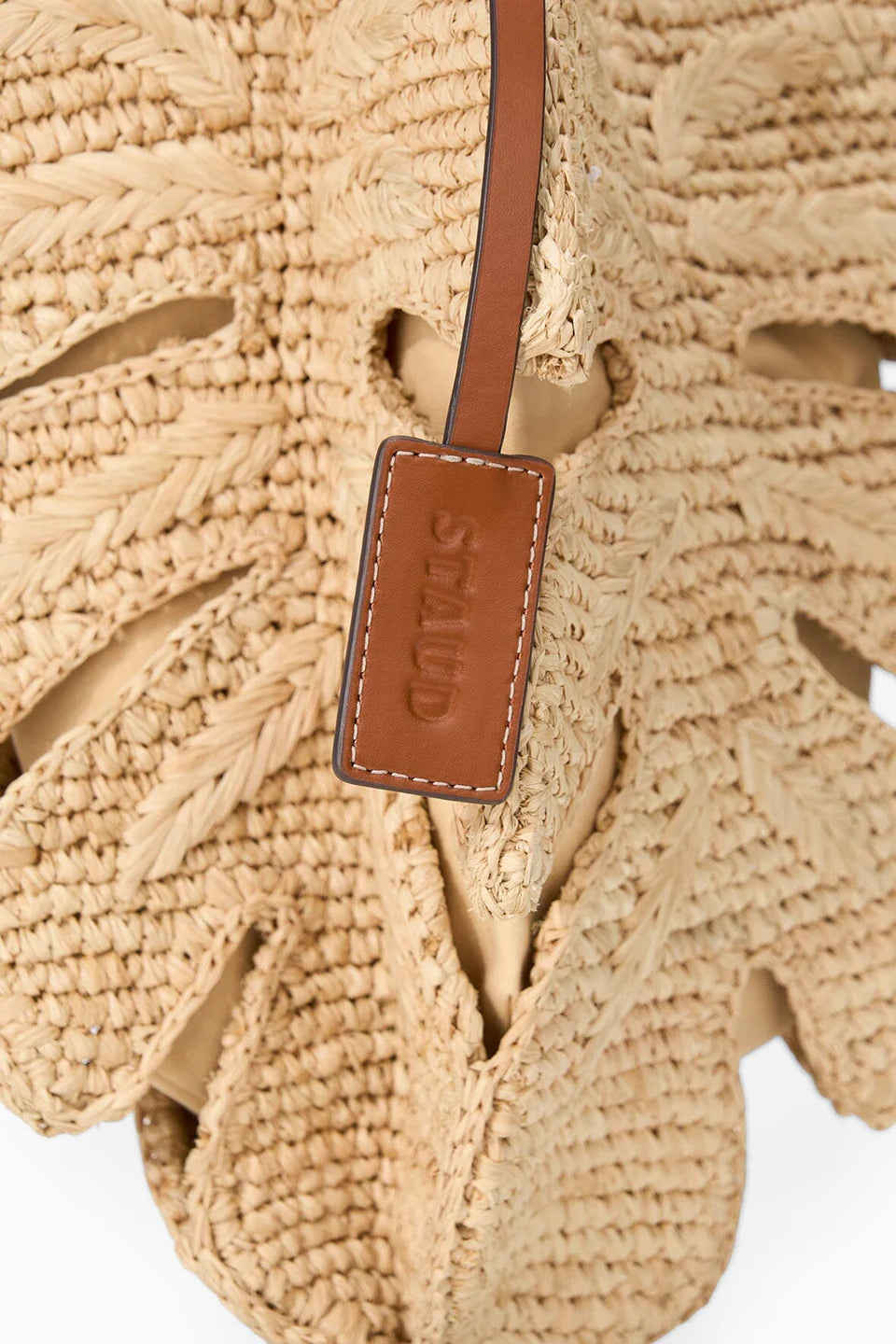 Close-up of the Staud Palm Raffia Bag, designed in a leaf shape with a brown leather tag embossed with the brand name.