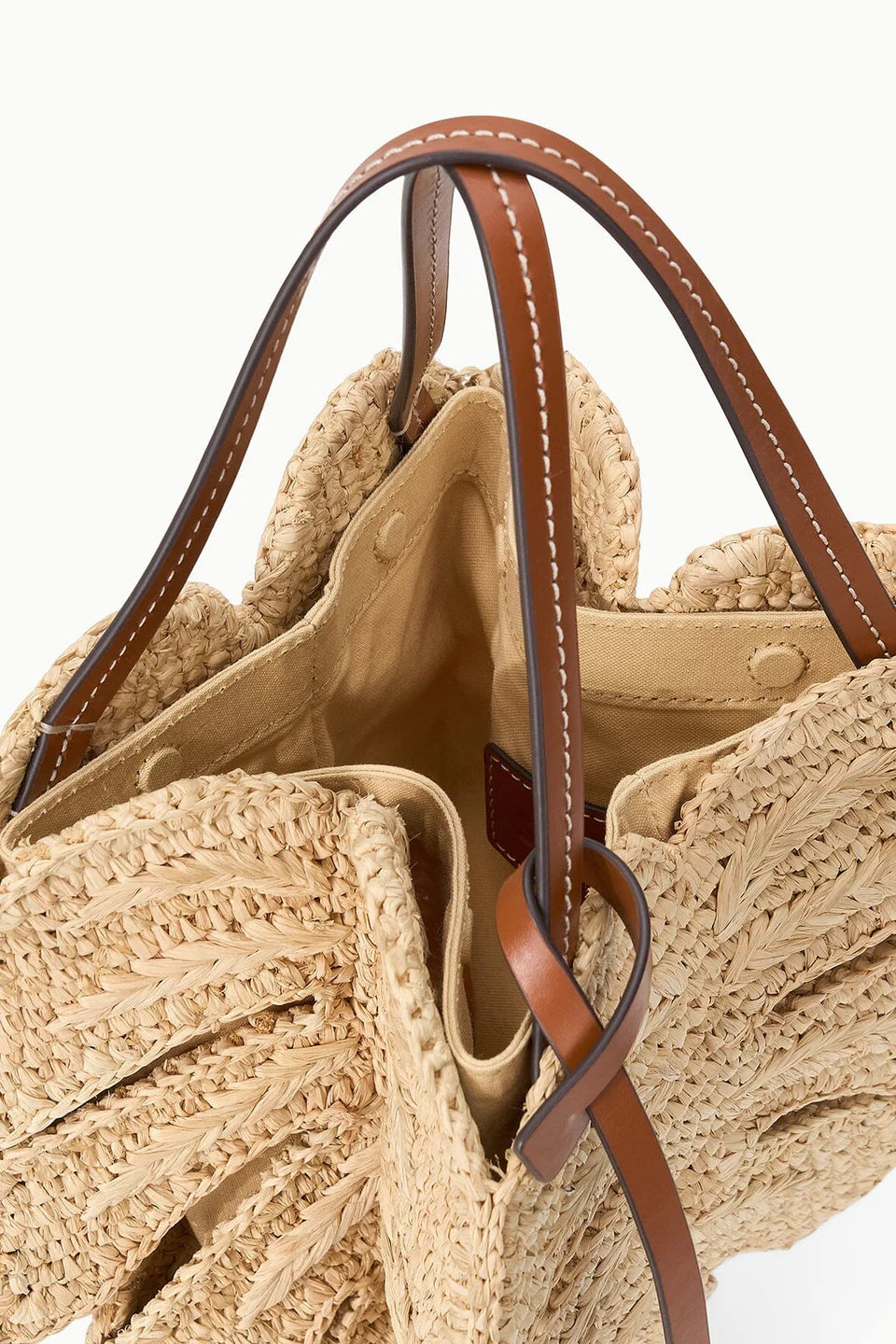 A close-up of the Staud Palm Raffia Bag features its spacious beige fabric-lined interior and open design with brown leather handles.