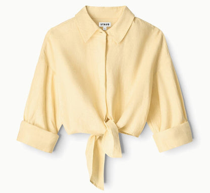 The Lisa Linen Shirt by Staud is a light yellow, long-sleeve cropped button-down with a collar, center front tie knot, and cuffed sleeves, shown against a white background.