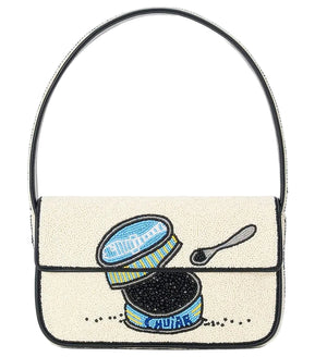 The Staud Tommy Beaded Bag is crafted from vegan leather and features hand-beading with an open caviar tin, small spoon, and caviar on a cream background, finished with a magnetic snap closure.