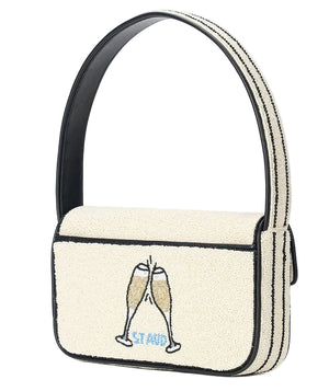 The Staud Tommy Beaded Bag is a cream-colored vegan leather shoulder bag with black trim, beaded with two clinking champagne glasses and "STAND" below, finished with a magnetic snap closure.