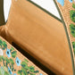 Explore the Staud Tommy Beaded Bag, an open handbag crafted from vegan leather featuring a hand-beaded floral design with blue and green flowers on a tan background. The interior is lined with elegant tan fabric, combining style with sustainability.