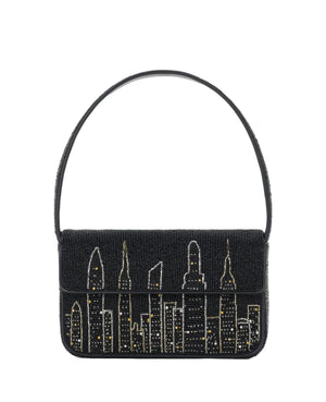 The Staud Tommy Beaded Bag is a black vegan leather handbag with a hand-beaded city skyline, gold and silver accents, a single shoulder strap, and a secure magnetic snap closure.