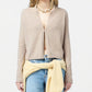 A person wearing the Guest in Residence Stealth Cardigan in beige over bare skin, paired with blue jeans, a yellow sweater tied at the waist, and a beaded necklace. The background is plain and light.