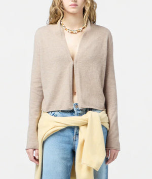 A person wearing the Guest in Residence Stealth Cardigan in beige over bare skin, paired with blue jeans, a yellow sweater tied at the waist, and a beaded necklace. The background is plain and light.
