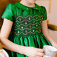 A young girl wears the Antoinette Baby Girls' and Girls' Stella Green Silk Dress as she sits on a chair, holding an heirloom white and gold teacup and saucer, as if celebrating a special occasion.