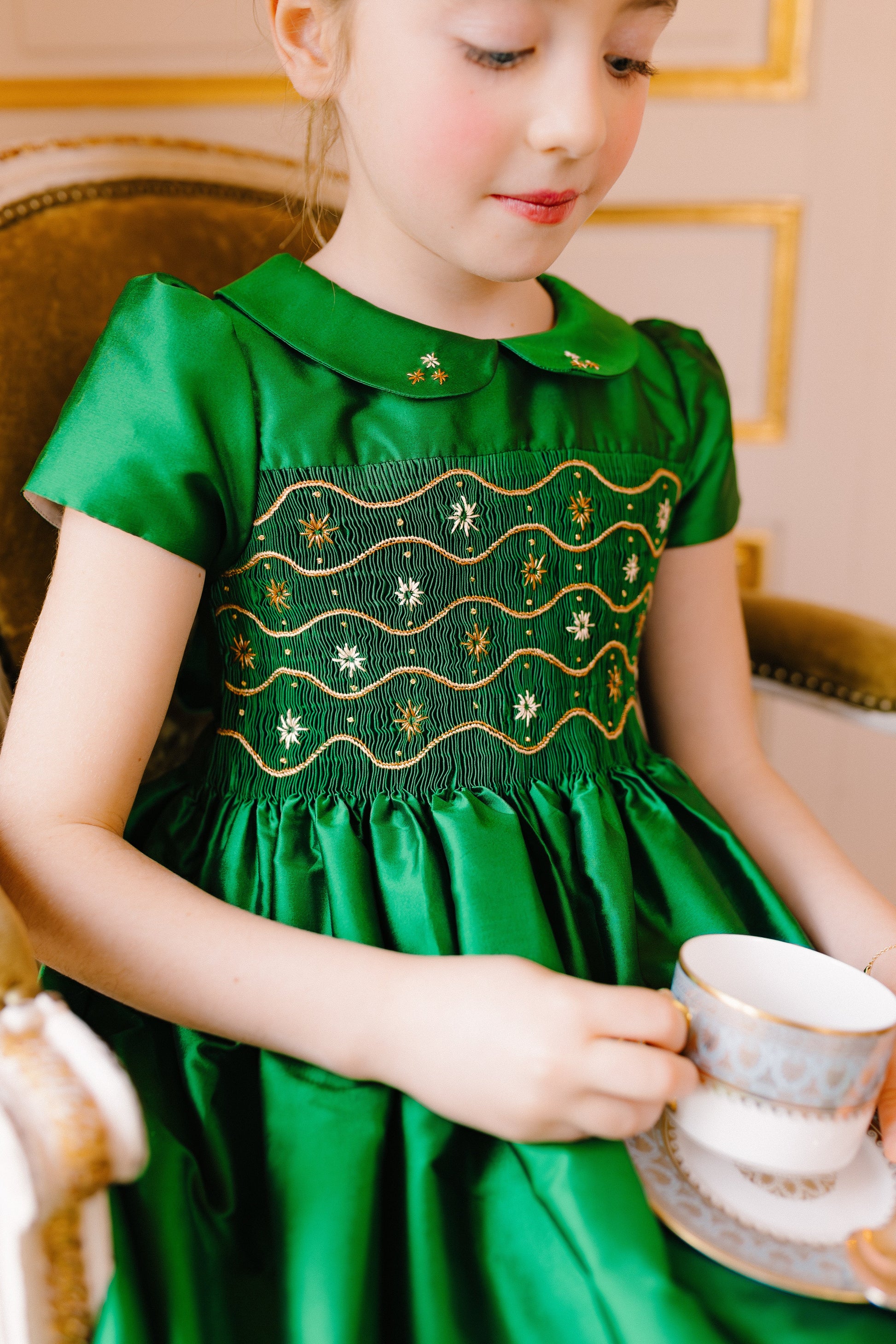 A young girl wears the Antoinette Baby Girls' and Girls' Stella Green Silk Dress as she sits on a chair, holding an heirloom white and gold teacup and saucer, as if celebrating a special occasion.
