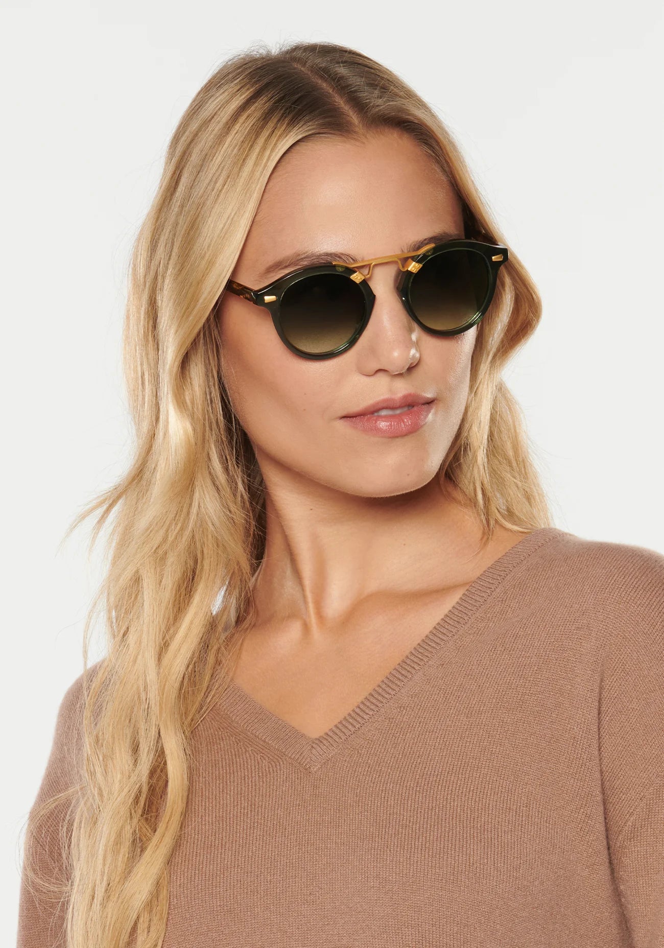 A woman with long blonde hair wears Krewe STL II sunglasses and a tan oversized V-neck sweater, standing against a plain light background.