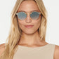 A woman with long blonde hair wears Krewe STL Nylon Mirrored Sunglasses featuring flat nylon lenses and a sleeveless beige top, facing forward against a plain white background.